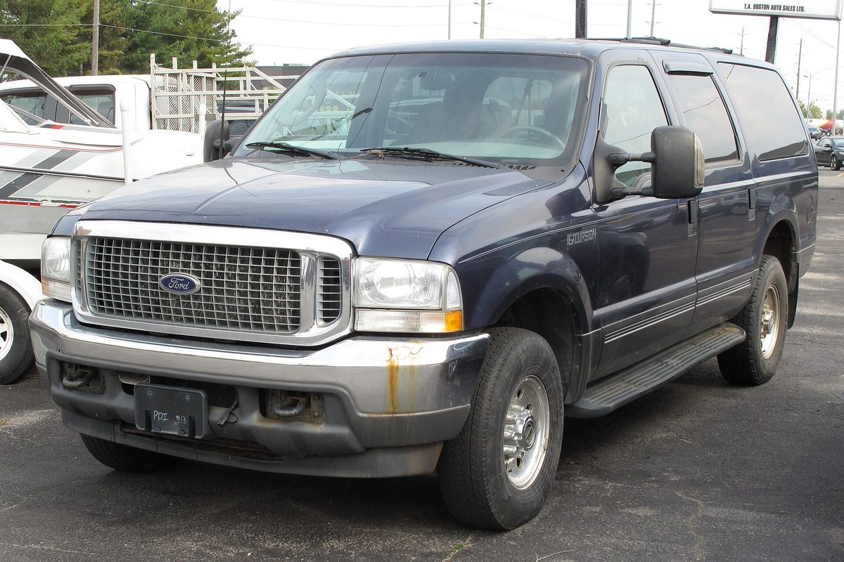 A dark blue 2003 Ford Excursion XLT with a chrome grille and rust stains on the front bumper, parked on a dealer lot