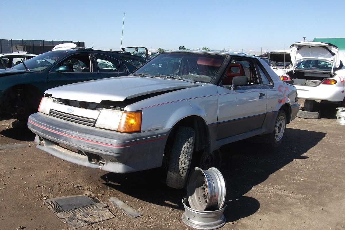 A silver late Ford Escort EXP with a battered front end in a dirt salvage yard, wheel rims stacked by its bumper