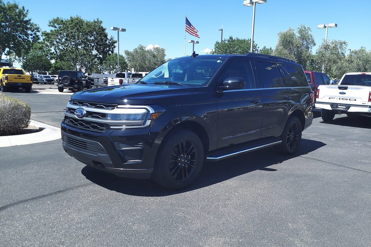 A black 2023 Ford Expedition XLT with black wheels on a dealer lot, an American flag and a white Ford pickup behind it