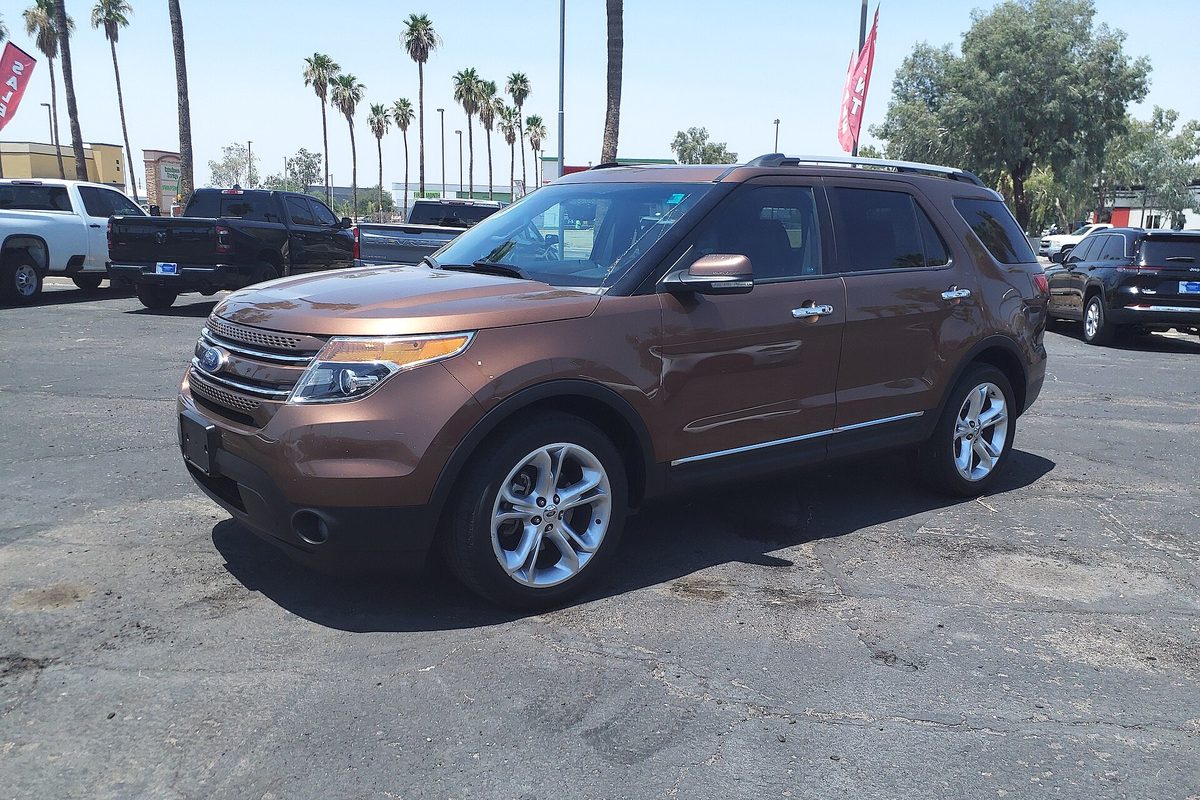 A bronze 2011 Ford Explorer Limited with chrome wheels on a sunny dealer lot, palm trees and pickups behind it