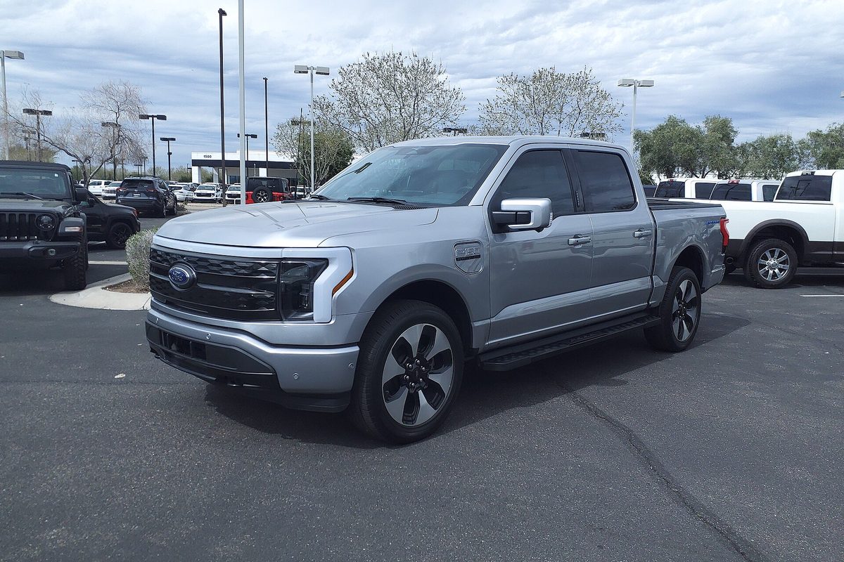 A silver 2023 Ford F-150 Lightning Platinum crew cab with black wheels, parked on a dealership lot under a cloudy sky