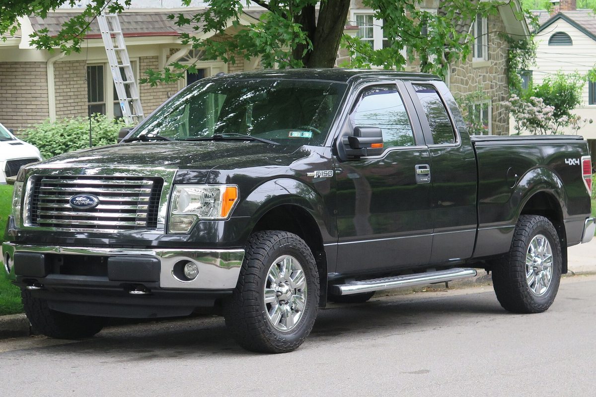 A black 2012 Ford F-150 XLT SuperCab 4x4 with chrome wheels and side steps, parked on a tree-lined residential street