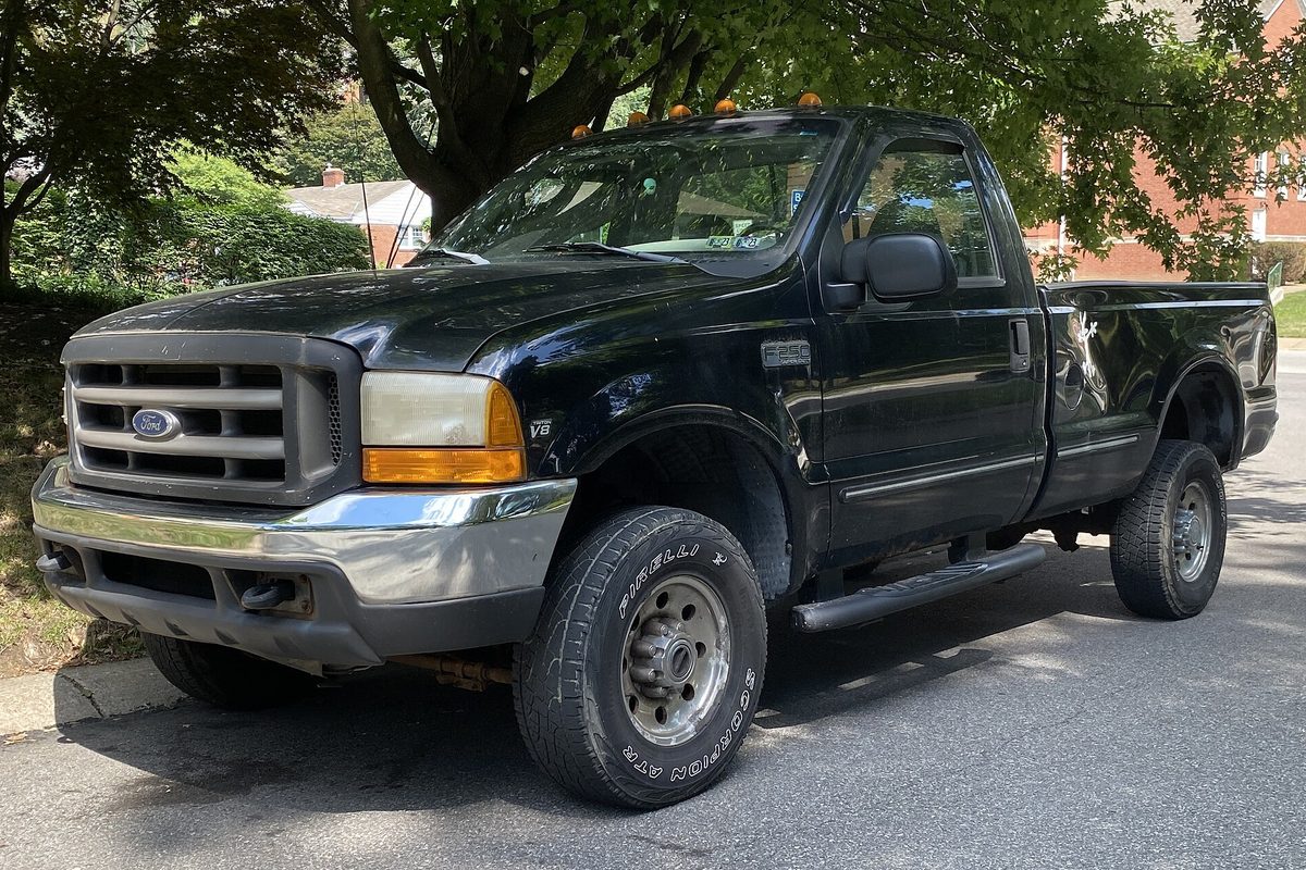 A black 1999 Ford F-250 Super Duty regular-cab pickup with amber cab-roof lights and a chrome front bumper, parked on a tree-lined residential street