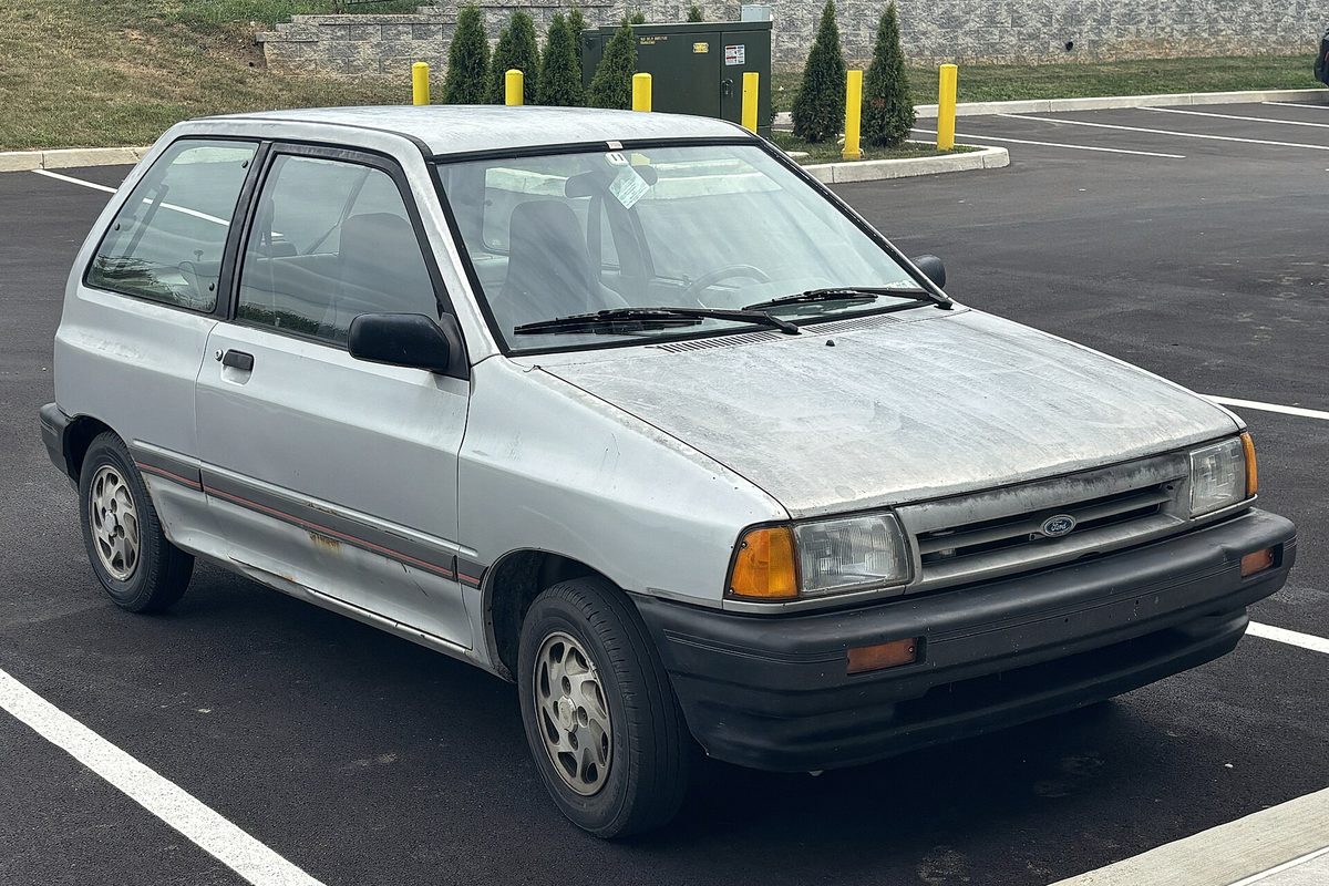 A silver 1989 Ford Festiva three-door hatchback with a sun-faded hood, parked alone in an empty lot