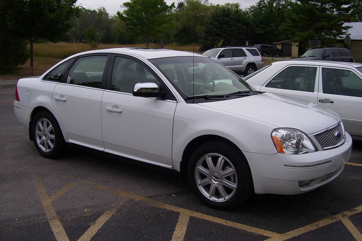 A white 2006 Ford Five Hundred sedan with chrome mirror caps and a mesh grille, parked between yellow lines in a lot beside another white car