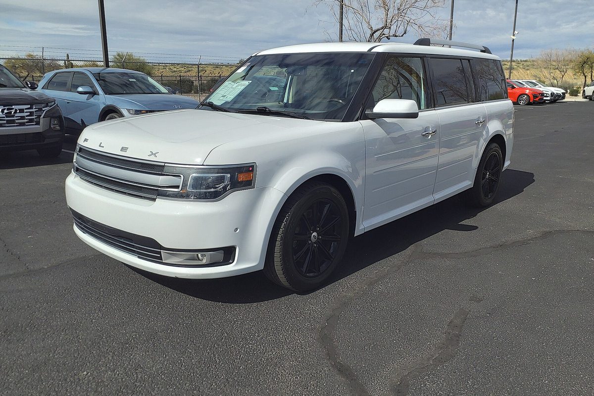 A white 2014 Ford Flex Limited with black wheels parked on a dealer lot, other cars and a chain-link fence behind it