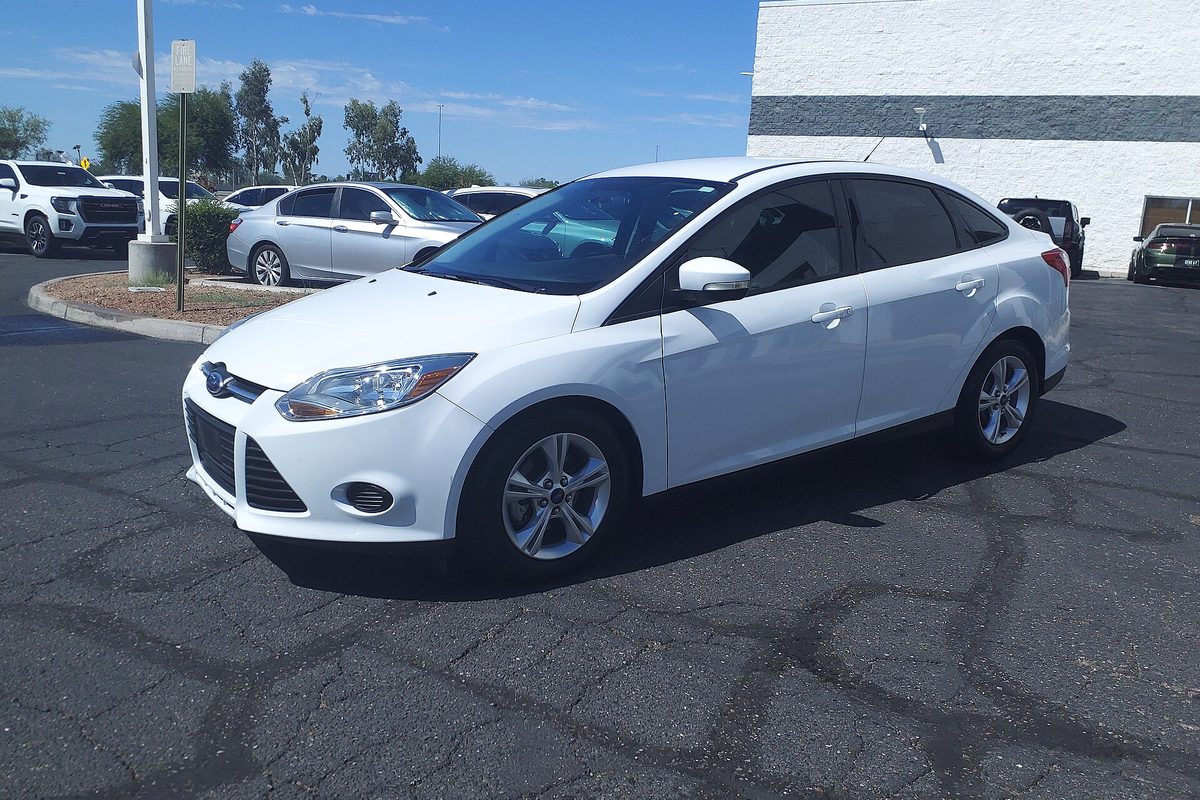 A white 2014 Ford Focus SE sedan parked on cracked asphalt at a dealer lot, with other cars behind it
