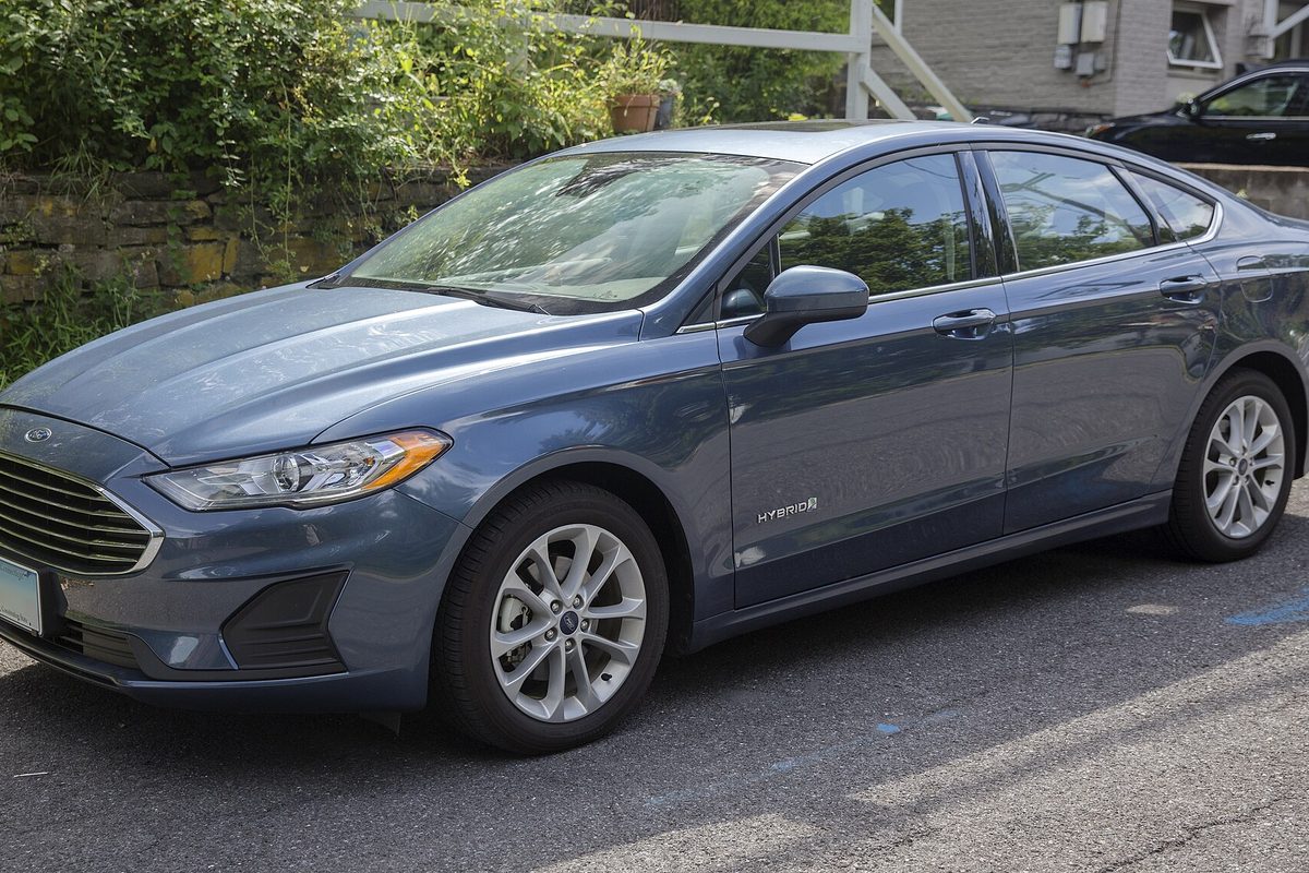 A blue-gray 2019 Ford Fusion Hybrid sedan parked on asphalt below a stone wall and green shrubs