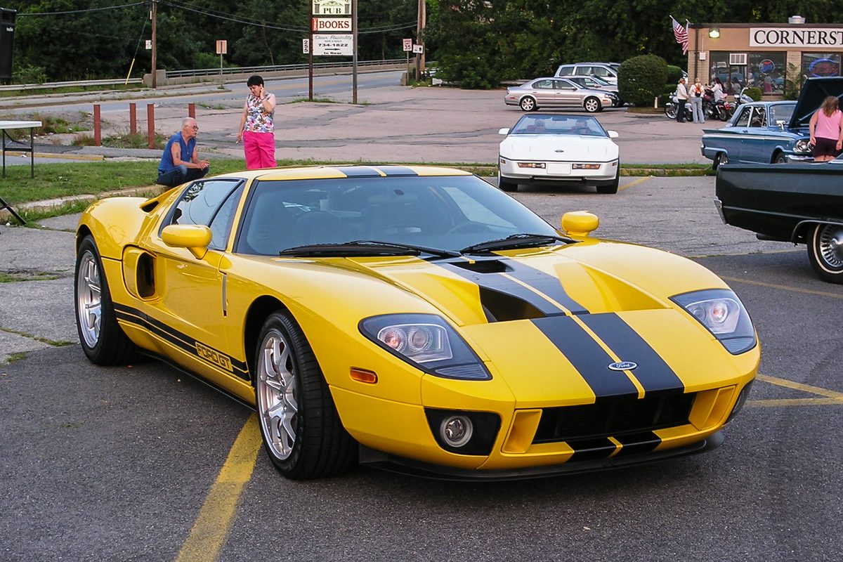 A yellow first-generation Ford GT with black stripes over the hood and roof at an evening car show in a parking lot