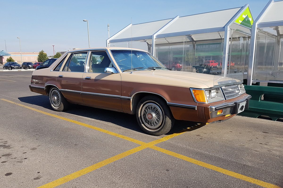 A two-tone brown and tan mid-1980s Ford LTD sedan with wire wheel covers, parked in a lot beside shopping cart shelters