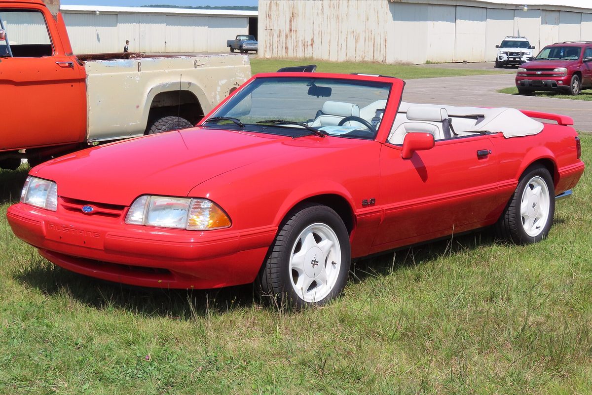 A red 1992 Ford Mustang LX 5.0 convertible with a white top and white wheels, parked on grass beside an old orange pickup at an airfield