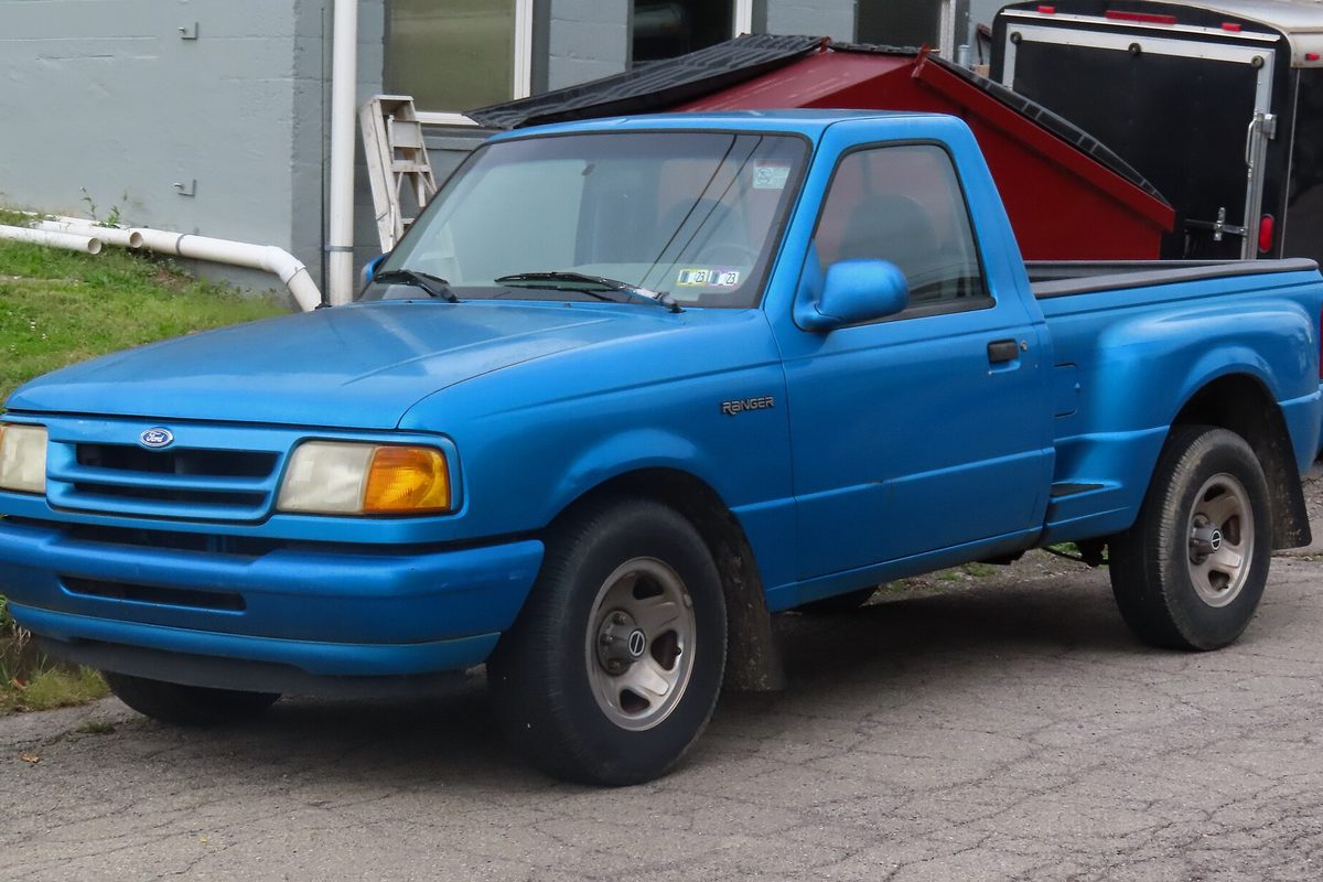 A bright blue 1994 Ford Ranger Splash regular-cab pickup parked on cracked pavement next to a small red shed with a black roof