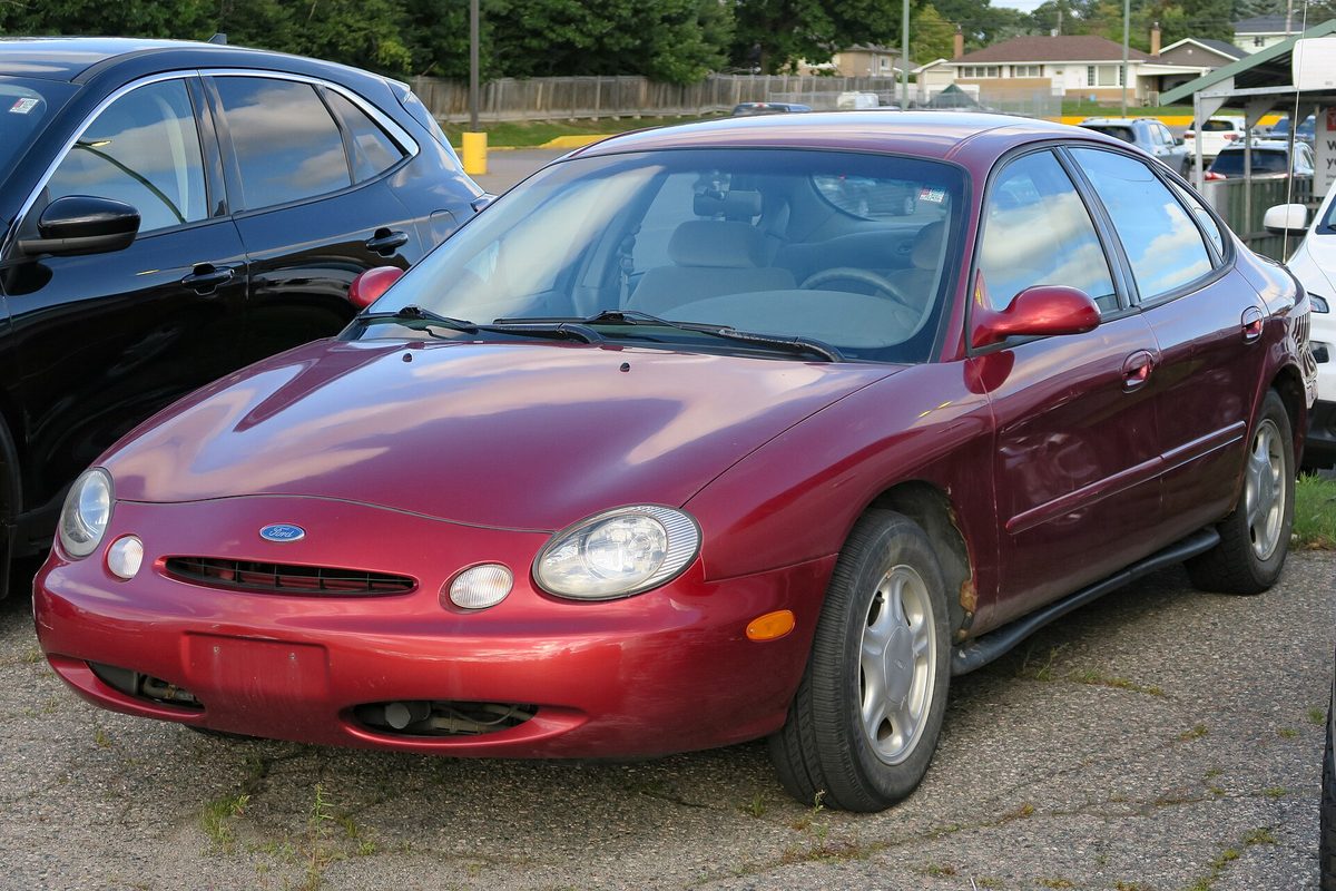 A dark red 1997 Ford Taurus GL sedan with oval headlights and a scuffed front bumper, parked on a gravel lot