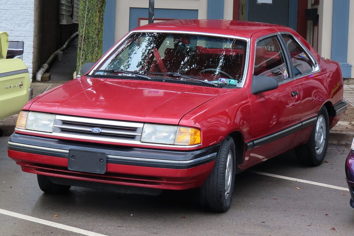 A red 1990 Ford Tempo GL two-door parked between white lines in a lot, with a pale green older car at the edge of the frame