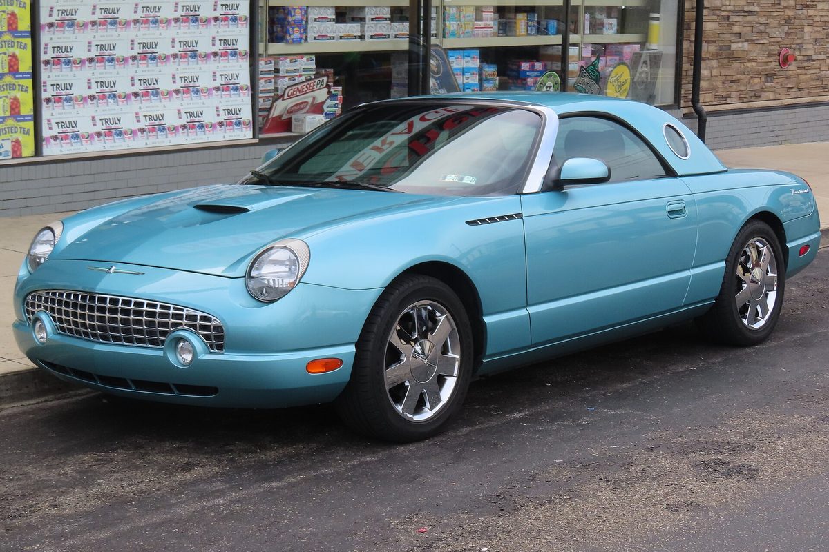 A light turquoise 2002 Ford Thunderbird convertible with its porthole hardtop on, parked at a curb in front of a store window
