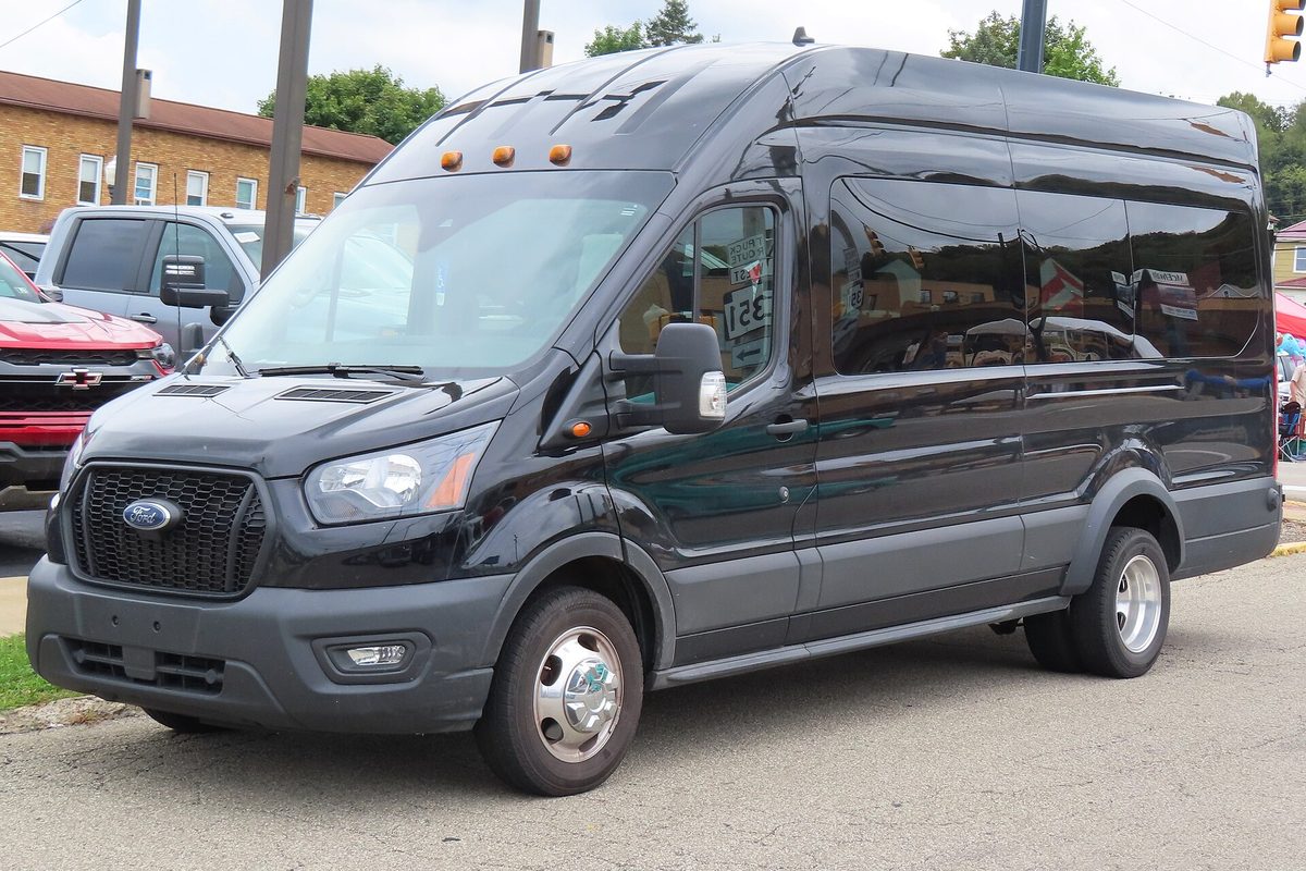 A black 2023 Ford Transit high-roof extended passenger van parked by a curb on a small-town street