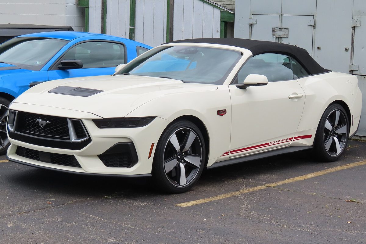 A white 2025 Ford Mustang GT convertible with its black top up and a red 60 Years stripe along the side, parked in a lot next to a blue car