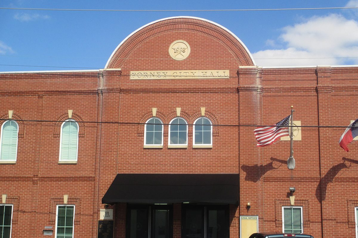 The red brick front of Forney City Hall with its arched roofline, a round date medallion, a black entrance awning and the U.S. and Texas flags