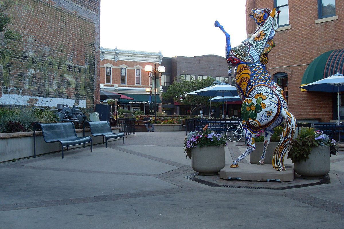 A tall mosaic horse sculpture covered in bright painted tiles in an Old Town Fort Collins plaza, with benches, planters and brick storefronts behind