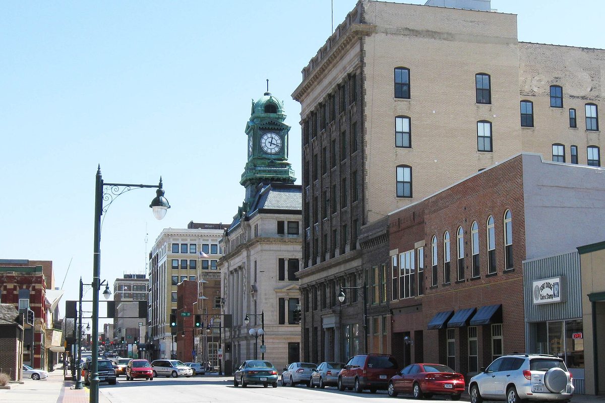 A downtown Fort Dodge street with a green copper clock tower on an old stone building, tall brick buildings and cars parked along the curb