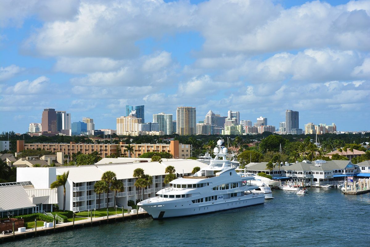 A large white yacht docked on a Fort Lauderdale waterway beside low white buildings, with the downtown skyline and palm trees behind under a cloudy blue sky
