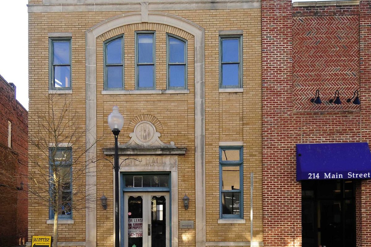 A tan brick two-story building with arched upper windows on Main Street in Fort Mill, next to a red brick storefront with a blue awning reading 214 Main Street