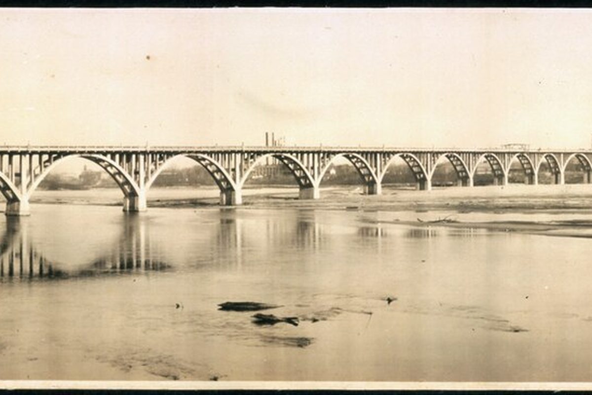An old sepia photograph of a long arched bridge crossing the Arkansas River at Fort Smith, with a smokestack on the far bank