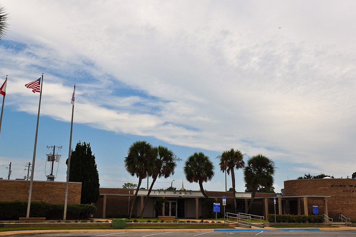 Fort Walton Beach City Hall, a low brick building behind a row of palm trees, with flagpoles out front under a sky of streaked clouds