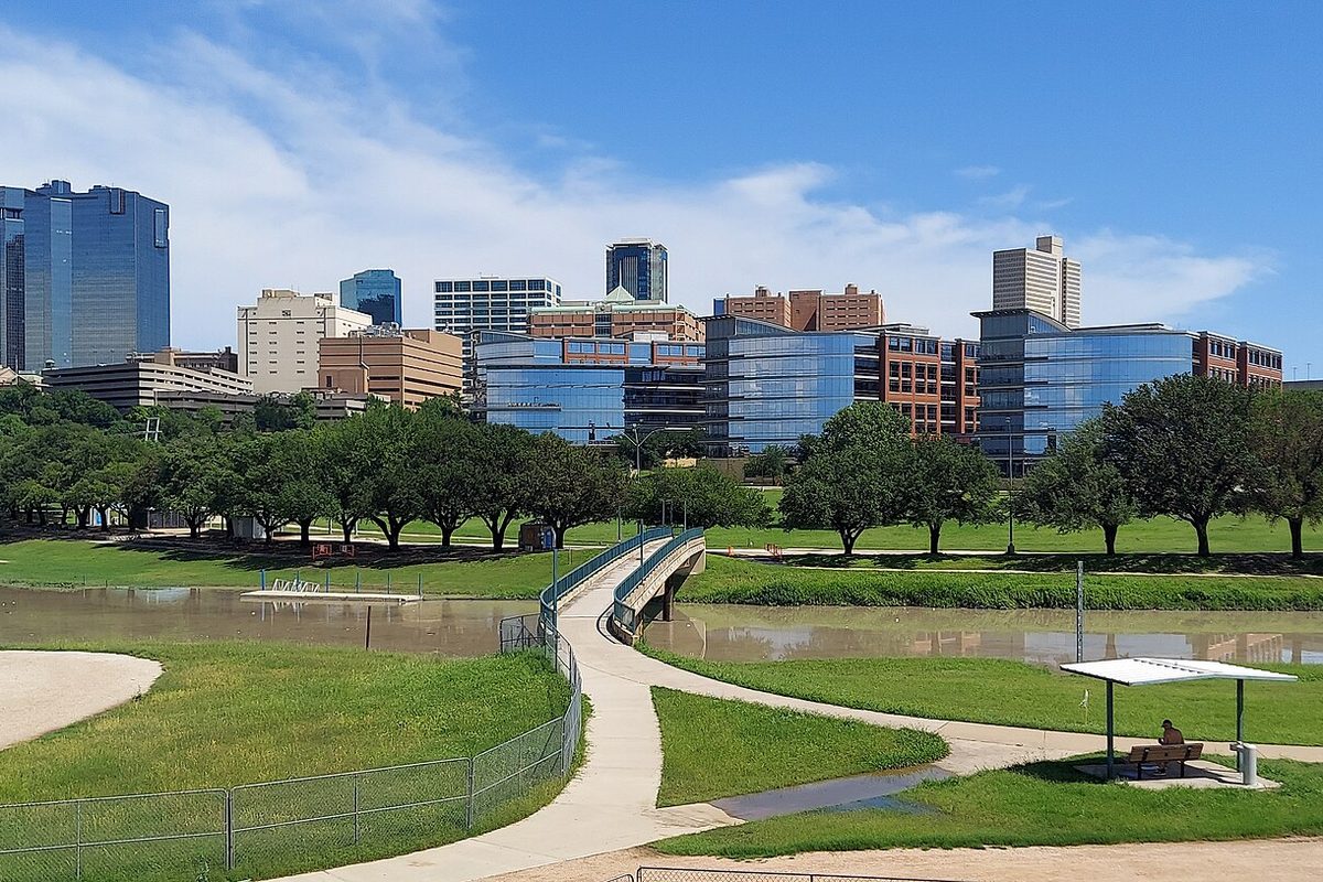 Downtown Fort Worth towers rising behind a row of trees, with a curving footbridge over a muddy channel and green lawns along the Trinity Trails