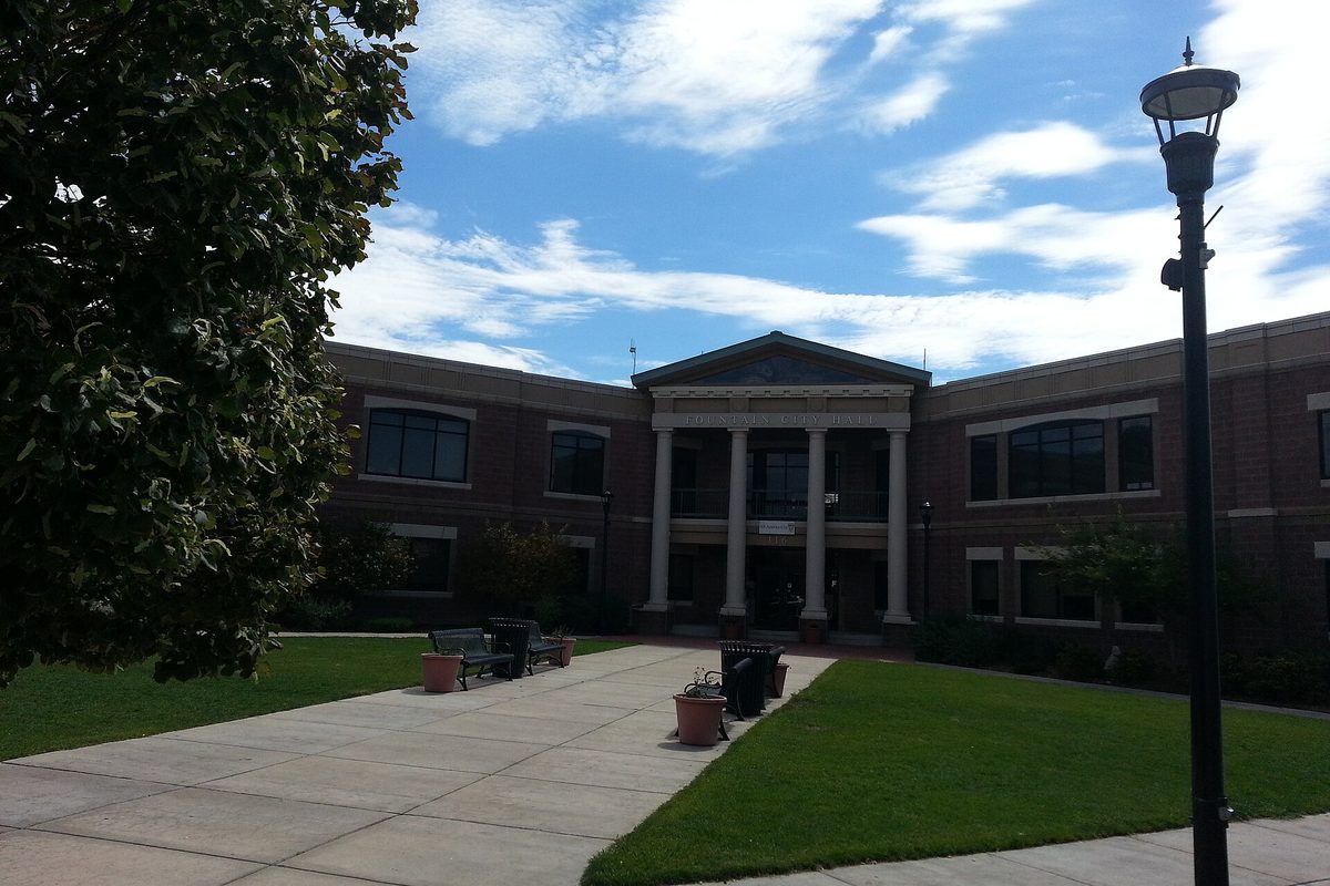 Fountain City Hall, a two-story brick building with a white-columned entrance, a lawn and a concrete walkway under a blue sky