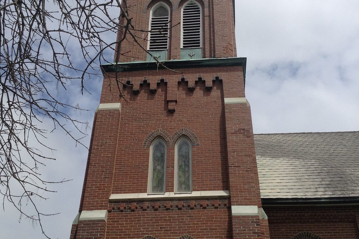 A red brick church tower in Frankfort with arched stained-glass windows and louvered belfry openings, seen through bare branches under a cloudy sky