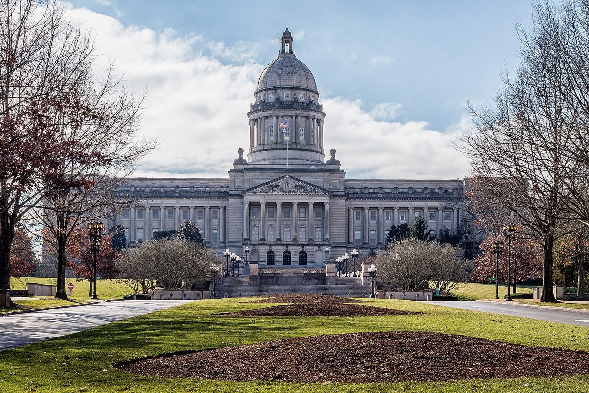 The domed Kentucky State Capitol in Frankfort seen across a lawn with mulched beds and bare trees under a cloudy sky