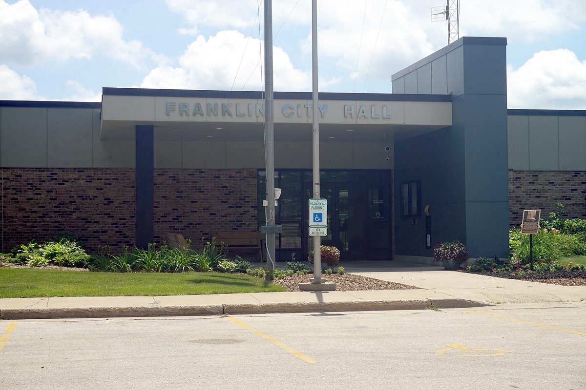The entrance of Franklin City Hall, with its name in raised letters above the doors, brick walls, a flagpole and flower beds