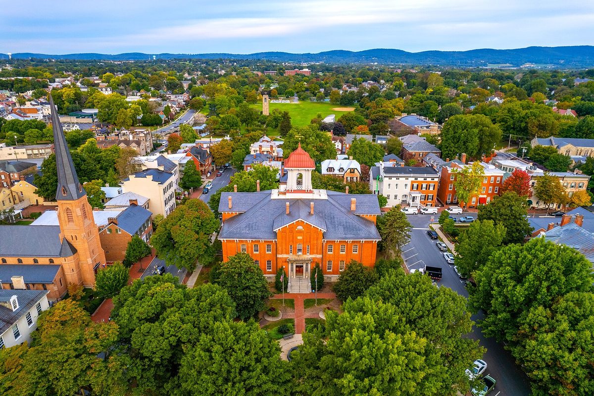 Aerial view of Frederick's red-brick City Hall with its white cupola, surrounded by tree-lined streets and church steeples, with mountains on the horizon