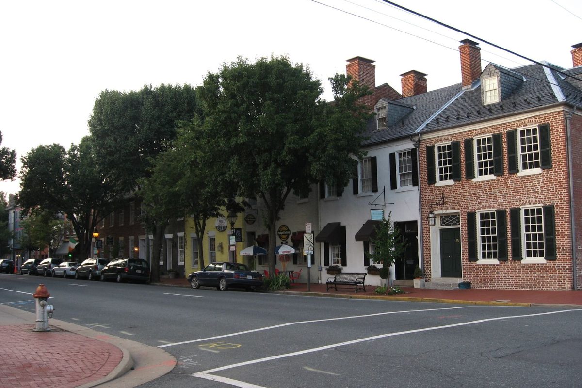 The corner of Caroline and Charlotte Streets in historic downtown Fredericksburg, with a red brick colonial house, painted storefronts, shade trees and parked cars