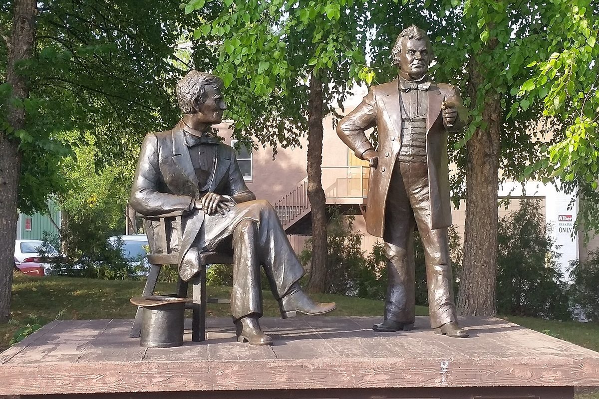 Bronze statues of Abraham Lincoln seated and Stephen Douglas standing on a stone platform under trees, the debate memorial in Freeport