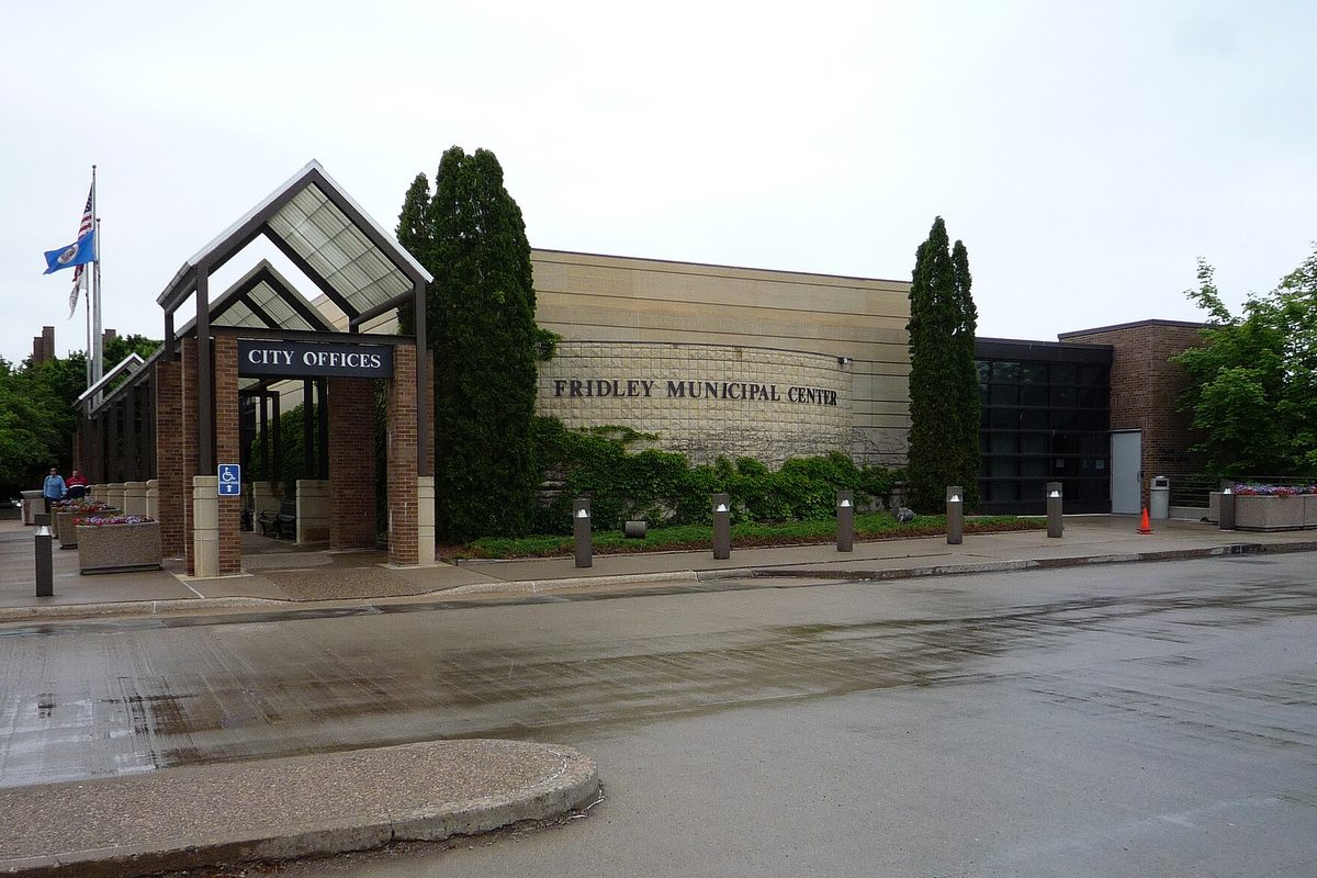Fridley Municipal Center, a low tan stone building with a glass-roofed City Offices entrance canopy, a flagpole with U.S. and Minnesota flags and tall evergreens, seen across a wet street