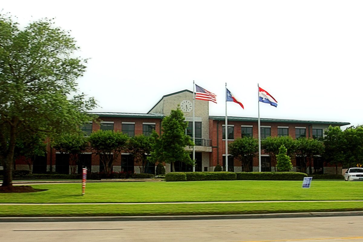 Friendswood City Hall, a two-story brick building with a clock above the entrance and three flags on poles, behind a wide green lawn