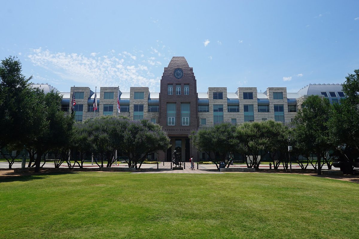 The George A. Purefoy Municipal Center in Frisco, a stone and brick building with a clock tower and flags, behind a wide lawn and trees