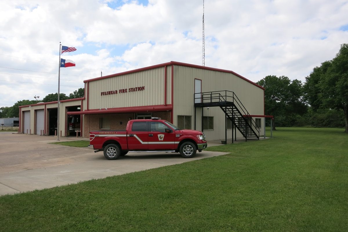 Fulshear Fire Station, a tan metal building with red trim and an outside stairway, with a red fire department pickup out front and U.S. and Texas flags flying