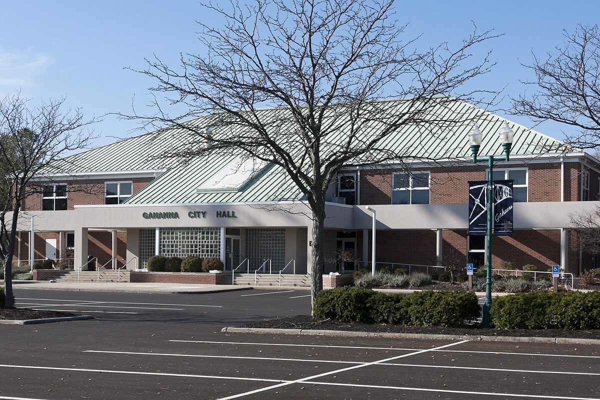 Gahanna City Hall, a brick building with a pale green metal roof and its name over the entrance, seen across an empty parking lot past a bare tree