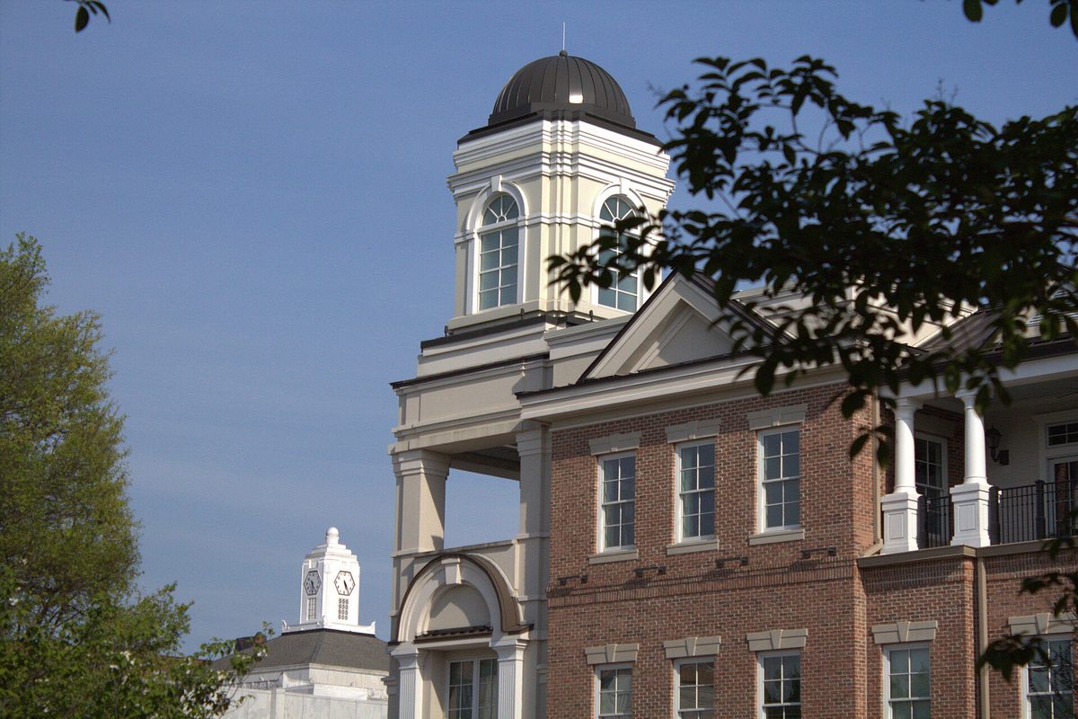 The domed tower and columned upper porch of a red-brick building in downtown Gainesville, with a white clock tower in the distance