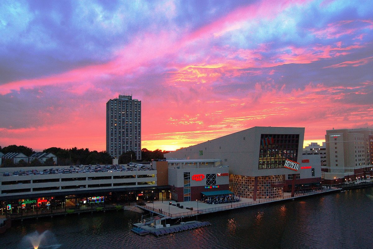 A tall apartment tower and a waterfront building with restaurants in Gaithersburg under a pink and purple sunset sky, with a lake and fountain in front