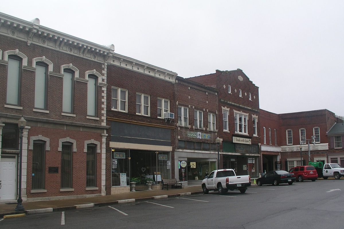 Two-story brick storefronts on the Gallatin town square with a white pickup and other cars parked along the curb under a gray sky