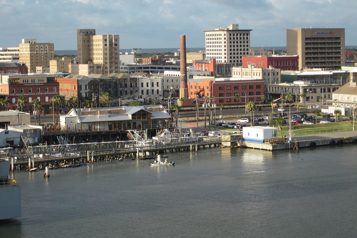 Downtown Galveston seen across the harbor, with brick and stone buildings, a tall brick smokestack, palm trees and piers along the waterfront