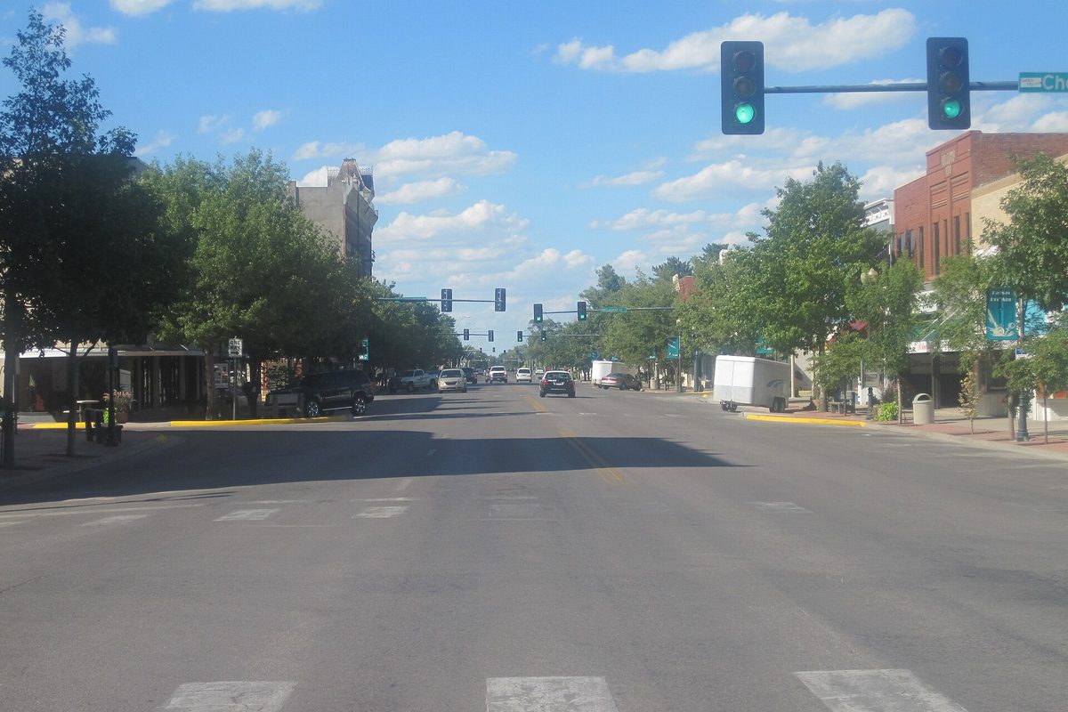 A wide downtown Garden City street with green traffic lights overhead, trees, parked cars and brick buildings under a blue sky