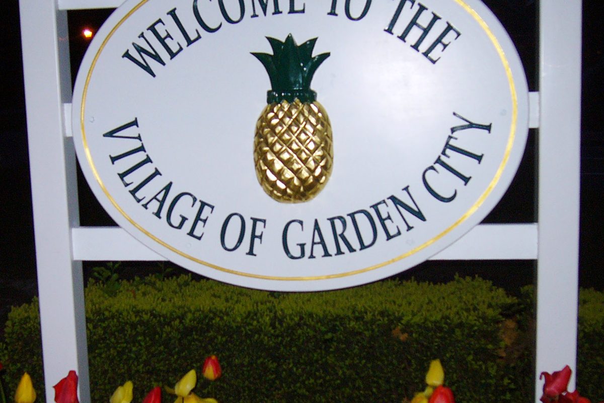 A white oval sign reading Welcome to the Village of Garden City, with a gold pineapple in the center, above a trimmed hedge and tulips