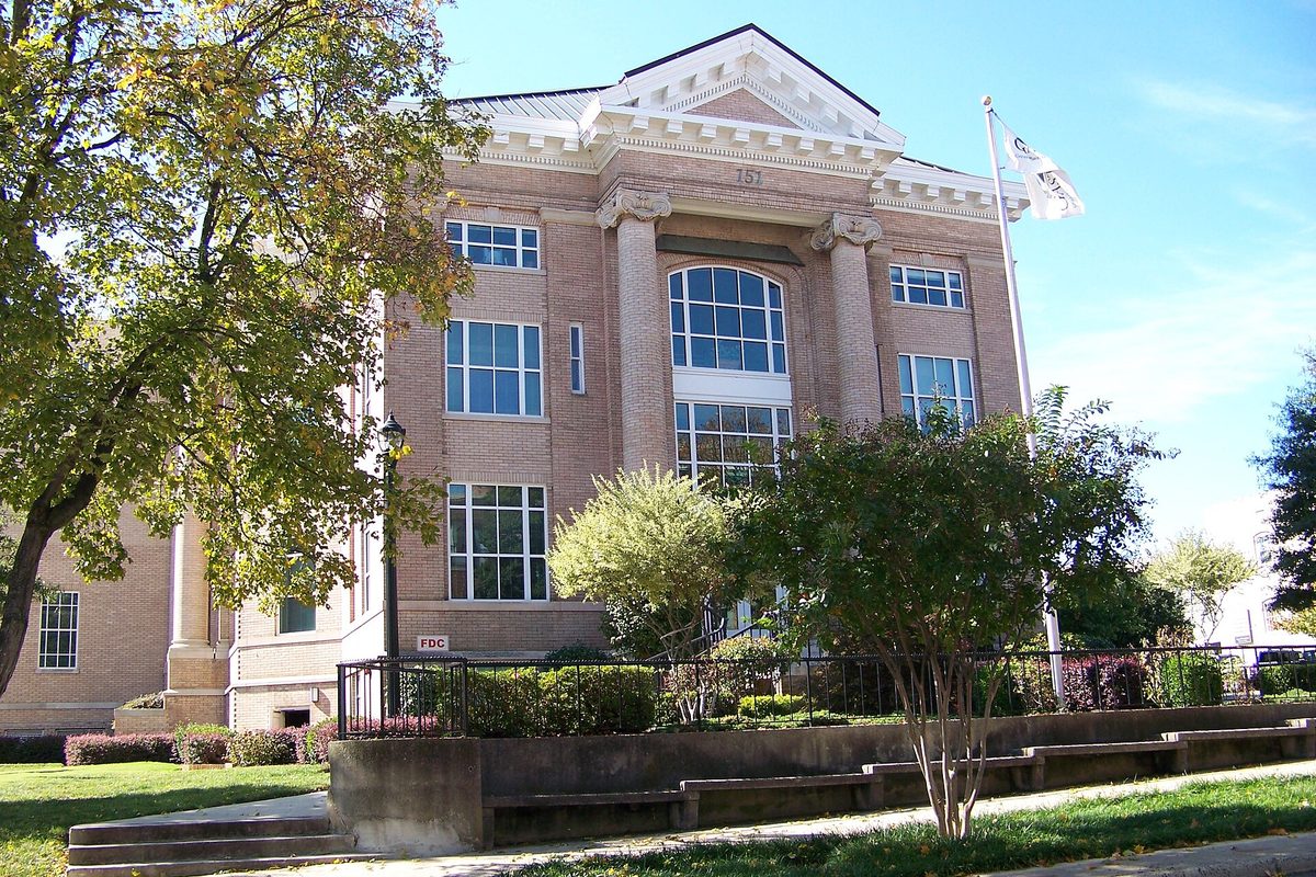 The historic 1910 Gaston County Courthouse in downtown Gastonia, a tan brick building with columns and a tall arched window, framed by trees on a sunny day