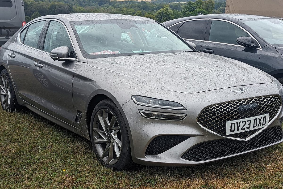 A silver 2023 Genesis G70 sedan, the facelifted version with split headlights, beaded with rain on a grass parking area