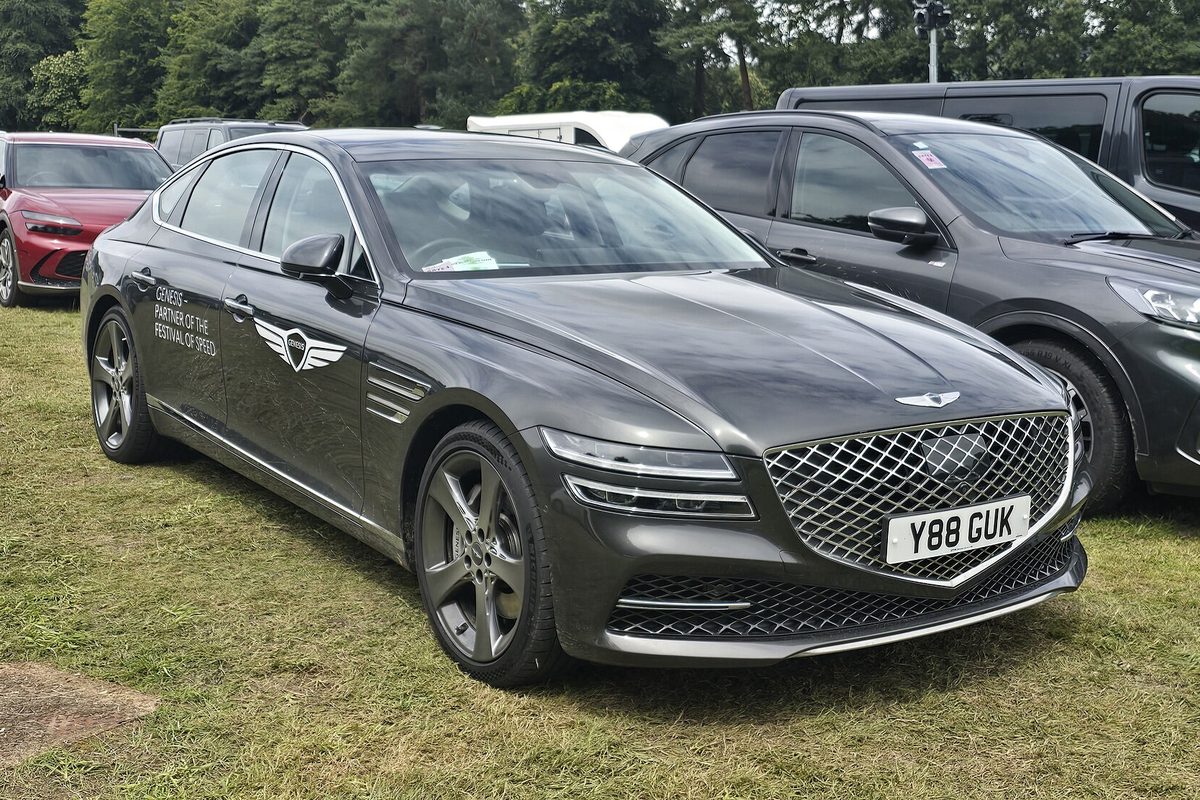 A dark gray second-generation Genesis G80 sedan with a wide grille, parked on grass at an outdoor car show
